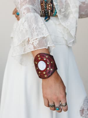 A close-up of a bold burgundy leather cuff bracelet with brass studs and a white stone centerpiece, worn against a white lace ruffle sleeve — layered with beaded bracelets and silver rings for that effortlessly collected boho wrist stack.