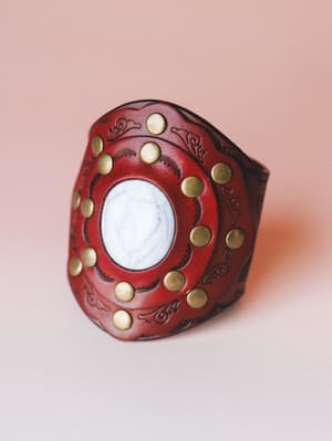 A bold, wide leather cuff bracelet in deep crimson, centered with a white howlite stone and ringed by brass studs — the kind of piece that makes a simple outfit feel like a whole story.