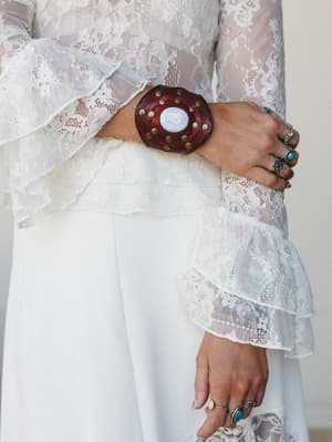 A close-up detail shot of a rich burgundy leather cuff bracelet with brass studs and a white stone center, worn over a white lace ruffle-sleeve top — the kind of accessory that does all the talking.
