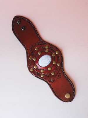 A wide tooled leather cuff bracelet laid flat against a blush background, centered by a white howlite stone surrounded by brass studs and intricate embossed scrollwork — the kind of piece that looks like it has a story.