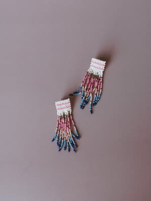 A pair of beaded fringe earrings laid flat on a dusty mauve surface — woven seed bead tops with a pink chevron pattern cascade into long multicolor strands that fade from white and pink through taupe into cobalt blue tips.