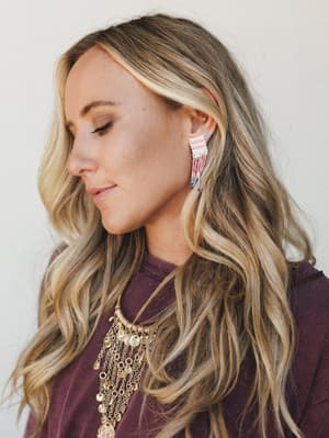 A close-up lifestyle shot showcasing beaded fringe drop earrings in pink and multicolor against a soft plum top, styled with a layered gold coin necklace — the whole look has that effortless boho-festival energy.