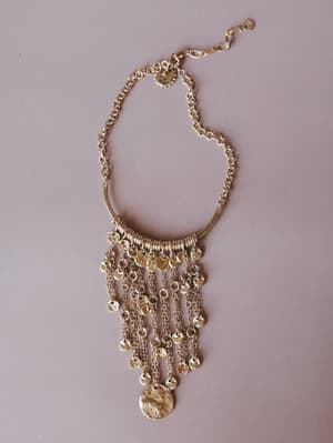 A gold statement necklace laid flat against a muted mauve background — the kind of piece that makes a plain white tee feel intentional, with cascading chain fringe, hammered coin charms, and a curved bar pendant that reads ancient-meets-everywhere.