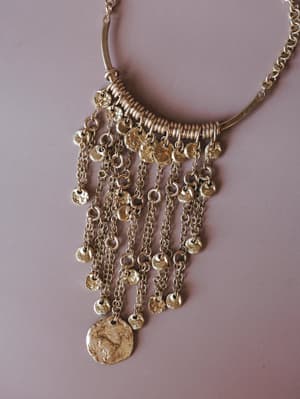 A gold statement necklace with a curved bar pendant wrapped in coiled rings, trailing multiple draped chains layered with hammered coin discs — ancient-feeling and effortlessly boho.