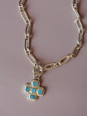 A chunky silver paperclip chain necklace with a detailed cross-shaped pendant set with four square turquoise stones, shot flat against a dusty mauve background — the kind of piece that makes a simple outfit feel like it has a backstory.