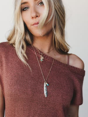 A close-up lifestyle shot focused on a layered gold necklace set with a turquoise stone pendant, worn against a dusty rose knit sweater — the kind of effortless boho styling that makes the jewelry feel personal, not precious.