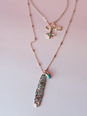 A layered gold necklace set styled against a soft blush background — the longer strand features an elongated filigree bar pendant with turquoise accents, while the shorter chain carries a star charm and small tag detail, the kind of piece that looks