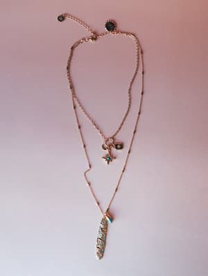 A layered gold chain necklace set laid flat against a soft pink surface — the shorter strand features a north star charm, crescent moon, and rectangular medallion, while the longer strand drops into an ornate filigree bar pendant with turquoise stone