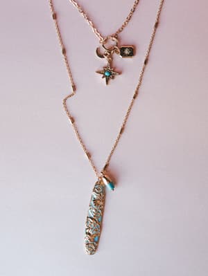 A layered gold necklace set styled flat against a soft blush background — the shorter strand clusters a crescent moon, north star, and dark enamel charm, while the longer chain drops into a filigree turquoise pendant with a beaded accent.