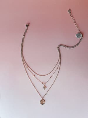 A silver layered necklace with three delicate chains — a beaded satellite chain, a dainty star charm, and a coin pendant — laid flat against a soft dusty rose gradient background.