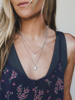 A close-up lifestyle shot showcasing layered silver necklaces — a dainty satellite chain, a north star pendant, and a moon medallion — styled against a navy floral tank top, with the focus squarely on the jewelry and how it sits.