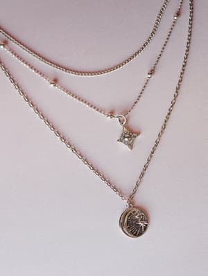 A layered silver necklace set arranged on a dusty rose surface, featuring a delicate satellite chain, a four-pointed star charm with crystal detail, and a longer chain with an engraved coin pendant etched with a crescent moon and starburst.