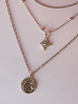 A close-up of a silver layered necklace set laid flat on a dusty pink surface — three delicate chains at varying lengths, one with a rhinestone star charm and one with an engraved coin medallion pendant.