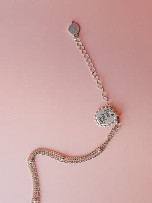 A silver layered chain necklace with a branded charm pendant and adjustable extender clasp, laid out against a soft blush pink background — delicate and easy to reach for every day.
