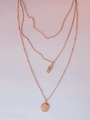 A delicate rose gold layered necklace set laid flat against a soft pink-to-lavender gradient background, featuring three chains of varying lengths — one plain curb chain, one satellite chain, and one with a small rhinestone bar pendant and a coin pen