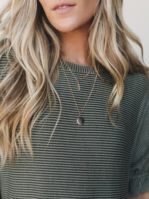A close-up shot of layered gold necklaces — a delicate chain, a small crystal pendant, and a dark moon disc pendant — worn over an olive striped ribbed tee, with soft wavy blonde hair framing the look.