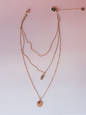 A delicate rose gold layered necklace set laid flat against a soft pink gradient background — three chains at varying lengths with a peace sign clasp, a small crystal bar charm, and a coin pendant with a crescent moon detail.