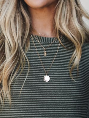 A close-up of layered gold necklaces — a dainty chain, a small crystal bar pendant, and a longer coin pendant — worn over a sage and charcoal striped ribbed top, with soft wavy blonde hair framing the shot.