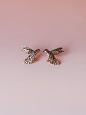 A pair of gold hummingbird stud earrings with intricate filigree detailing, shot flat against a soft dusty rose gradient background — delicate and a little magical.