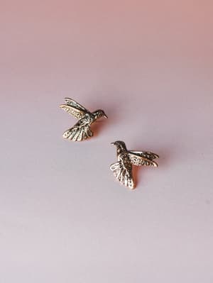 A pair of gold hummingbird stud earrings with intricate filigree detailing, laid against a soft blush-to-lavender gradient background — the kind of little treasure you'd notice on someone and immediately ask about.