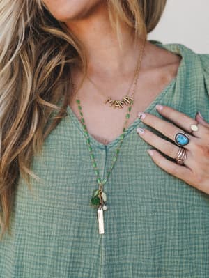 A close-up lifestyle shot layering a gold 'MAMA' letter necklace with a long green enamel bead chain and gold bar cluster pendant — styled against a sage textured top and paired with a turquoise stone ring for that effortless boho-collected look.