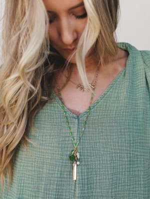 A close-up styled shot of layered necklaces against a sage green textured top — the long green enamel chain with mixed charms and gold bar pendant paired with a dainty gold letter necklace feels personal and quietly expressive.