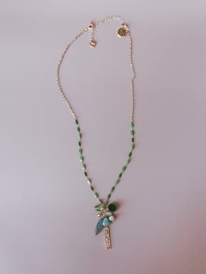 A delicate gold chain necklace with green enamel links and a mixed charm pendant cluster — featuring stone drops, a faceted crystal, and a textured gold bar — photographed flat on a soft blush surface.