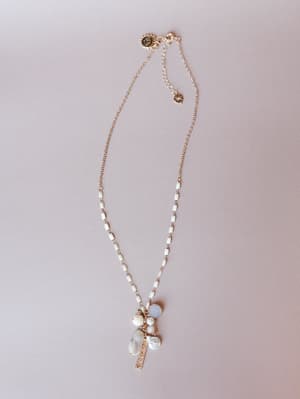 A delicate gold chain necklace with white enamel links and a clustered pendant of baroque pearls, mixed stone charms, and a pavé bar drop, laid flat against a soft blush-gray background.