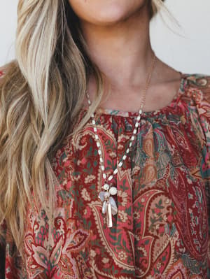 A close-up detail shot of a long pendant necklace layered over a rich paisley blouse — the white bead chain and mother-of-pearl cluster pendant feel earthy and collected, like something you'd find at a market and never take off.