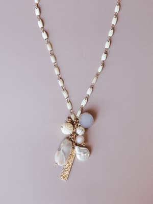 A long white enamel and gold chain necklace with a clustered pendant of baroque pearls, howlite beads, a frosted stone, and an engraved gold bar charm — laid flat on a soft mauve background.