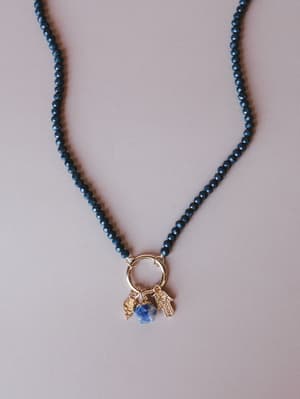 A deep navy faceted bead necklace laid flat, centered by a gold ring pendant clustered with three charms — a hamsa hand, a raw sodalite stone, and a small coin charm — all sitting against a soft mauve-grey background.