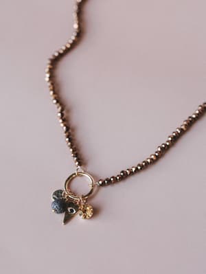 A close-up of a bronze-beaded necklace with a clustered charm pendant — a gold ring connector holds a lava bead, a gold heart charm, and a faceted amber crystal drop, all laid flat on a dusty rose surface.