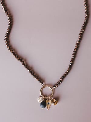 A close-up of a faceted bronze crystal bead necklace with a gold ring pendant clustered with mixed charms — a hammered coin, a lava stone bead, a gold heart with an evil eye detail, and an amber crystal drop — shot flat against a soft mauve backgroun