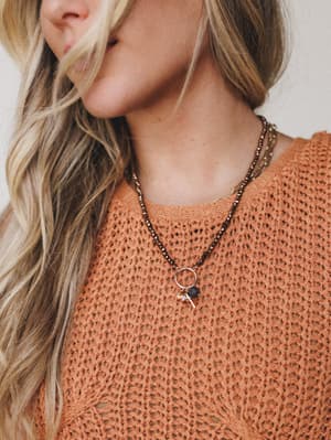 A close-up lifestyle shot of layered beaded necklaces styled against a rust orange open-knit sweater — warm, earthy, and effortlessly personal.