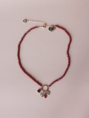A delicate red crystal beaded necklace laid flat on a blush surface, centered by a gold charm ring holding a cluster of mixed charms — a heart, a red teardrop gem, and a clear crystal drop — with an adjustable gold extension chain.