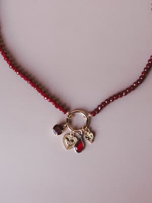 A close-up of a deep red faceted bead necklace with a gold ring clasp pendant cluster featuring a heart charm, teardrop red crystal, and small stone drop — the kind of piece that feels like a secret.