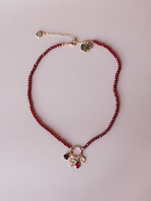 A deep red faceted crystal bead necklace laid flat on a muted mauve surface, centered by a gold ring charm cluster with a heart pendant, teardrop gem, and coin charm — the kind of piece that feels like it has a story.