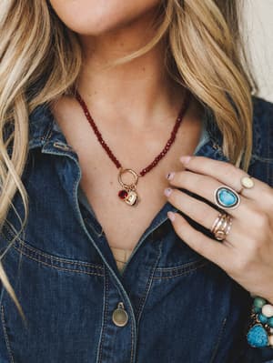 A close-up jewelry shot layering a deep red beaded necklace with a gold charm pendant against a denim shirt, styled with a stacked mix of turquoise, stone, and gold rings — the kind of accessory moment that looks effortless but is absolutely intentio