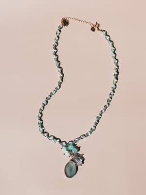A beaded statement necklace laid flat on a warm blush background, featuring translucent aqua glass beads and a cluster pendant with a marbled stone centerpiece — the kind of piece that makes a simple white dress feel like a whole moment.