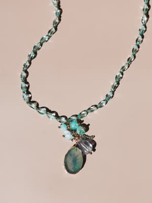 A beaded statement necklace with translucent aqua glass beads and a cluster pendant featuring a gold-edged stone slice, laid flat against a warm blush surface in directional sunlight.