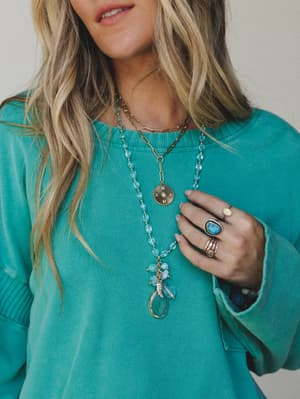 A close-up lifestyle shot showing layered gold necklaces — a long beaded blue stone strand with a crystal drop pendant and a chunky paperclip chain with a coin charm — styled over a washed teal crewneck sweatshirt, with stacked turquoise and stone ri