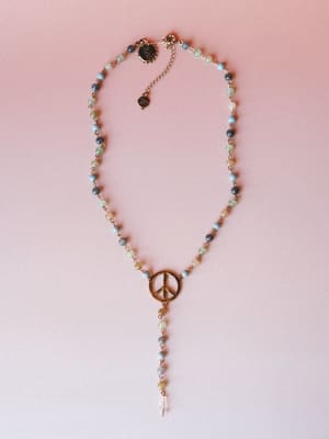 A delicate beaded lariat necklace laid flat on a soft pink surface — mixed stone beads in muted blues and greens lead down to a gold peace sign charm with a dangling crystal drop, giving it that free-spirited, quietly sentimental energy.