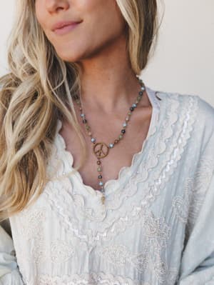 A close-up lifestyle shot of a beaded lariat necklace with a brass peace sign pendant, layered over a soft blue embroidered top — the kind of piece that feels like it has a story.