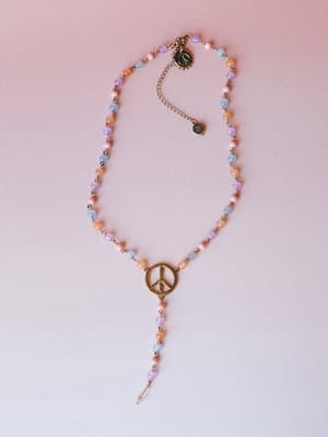 A lariat-style beaded necklace with a brass peace sign pendant, strung with small pastel beads in lavender, peach, and blue, laid flat against a soft pink-to-white gradient background.