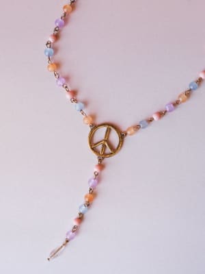 A delicate lariat-style beaded necklace laid flat on a soft blush surface — multicolor pastel beads linked by antique brass chain meet at a worn gold peace sign pendant, with a tiny crystal drop finishing the tail.