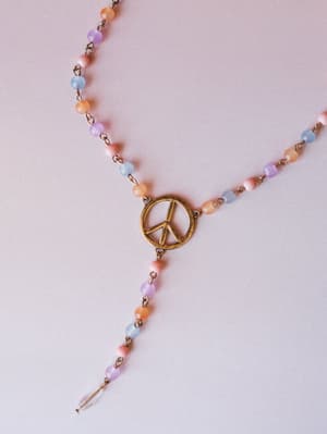 A delicate lariat-style beaded necklace laid flat on a soft blush surface — multicolor pastel beads linked by antique brass chain meet at a worn gold peace sign pendant, with a tiny crystal drop finishing the tail.