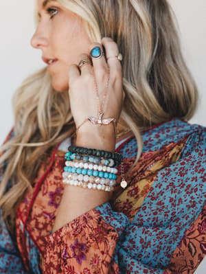 A close-up lifestyle shot of a model's hand and wrist stacked with beaded bracelets and a rose gold bird hand chain, rings layered across her fingers, posed against a softly blurred floral patchwork top — all the jewelry energy, none of the fuss.