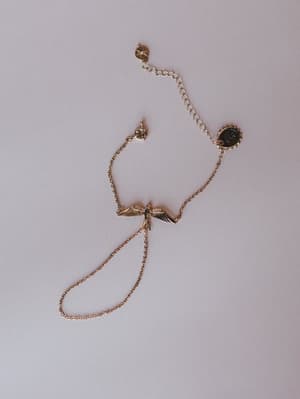 A gold hand chain bracelet laid flat on a soft grey surface, featuring a delicate bird-with-wings centerpiece charm, a small oval coin charm, a starburst charm, and a finger loop — the kind of piece that makes a simple outfit feel like it has a story