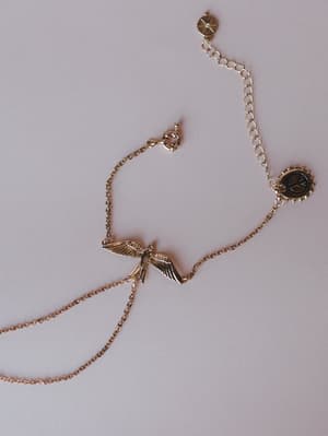 A delicate gold hand chain bracelet laid flat on a soft lavender-gray surface, centered by a detailed bird-in-flight pendant with layered chain loops, a small coin charm, and an extender chain — the kind of piece that feels like it has a story.