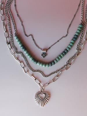 A layered silver necklace set styled flat against a dusty rose background — mixing a paperclip chain, curb chain, green beaded strand, and a delicate chain with a flower charm, all anchored by a sunburst heart pendant.